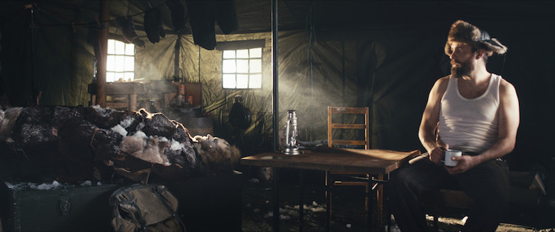 A man in a fur trimmed cat sits in a cabin and observes a man on a bed covered in ice